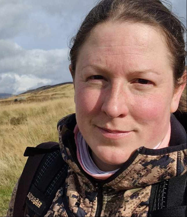A woman wearing a camouflage jacket with a backpack in a field